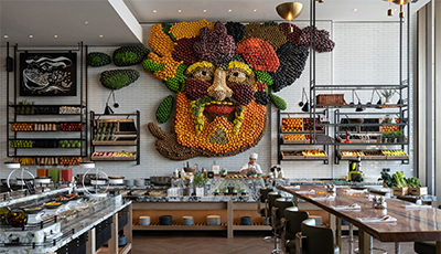 Restaurant interior with a buffet counter and a large wall artwork of a face made from fruit and vegetables