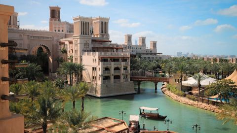 Sand-coloured wind-tower buildings and palm trees along a turquoise canal with a wooden abra moored at Madinat Jumeirah