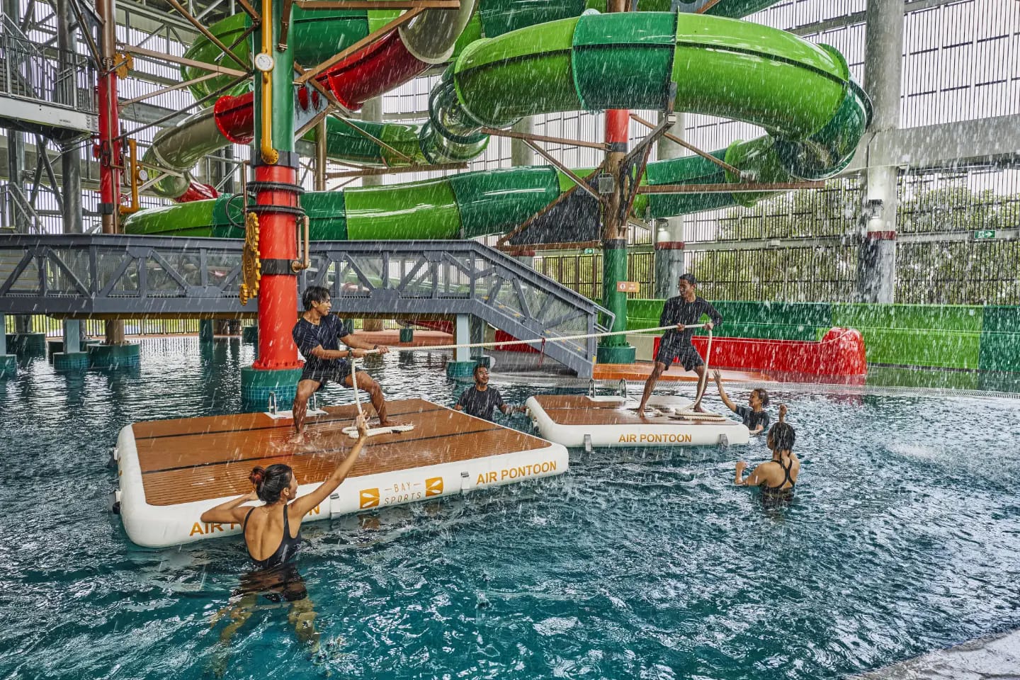 Visitors playing tug of war on floating air pontoons beside green water slides at Aqua Adventure