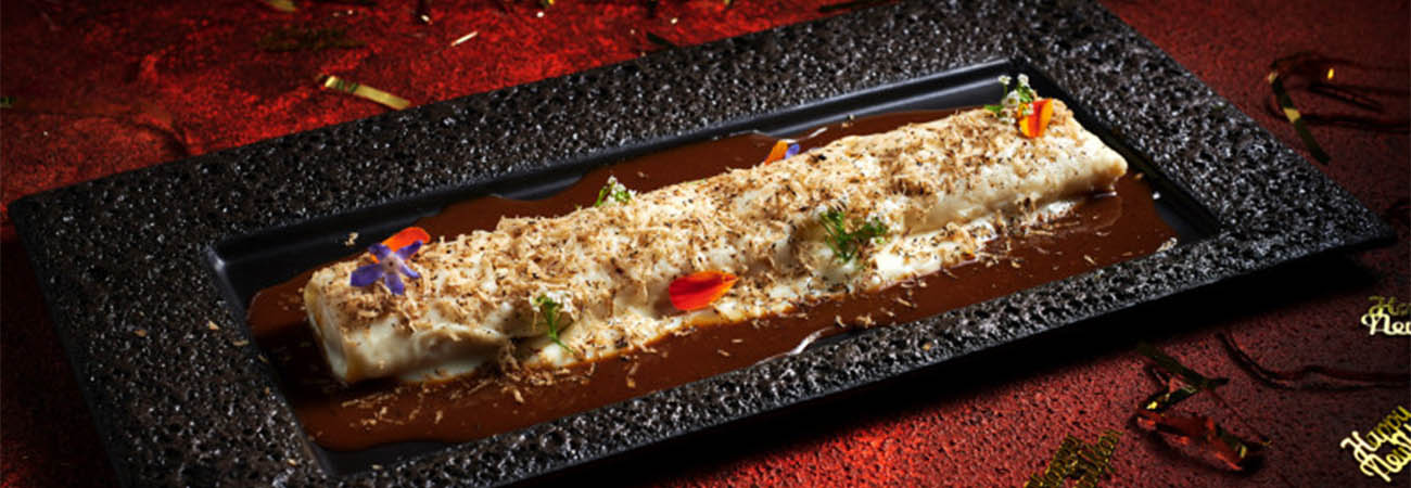 Plated crêpe roll topped with shavings, herbs and edible flowers on a black textured dish, with New Year confetti around it